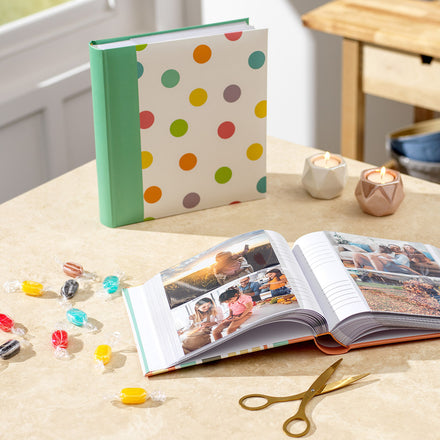 A colorful polka dot photo album with a teal green spine is displayed next to an open album showing printed photos, alongside scattered candies and a pair of scissors.